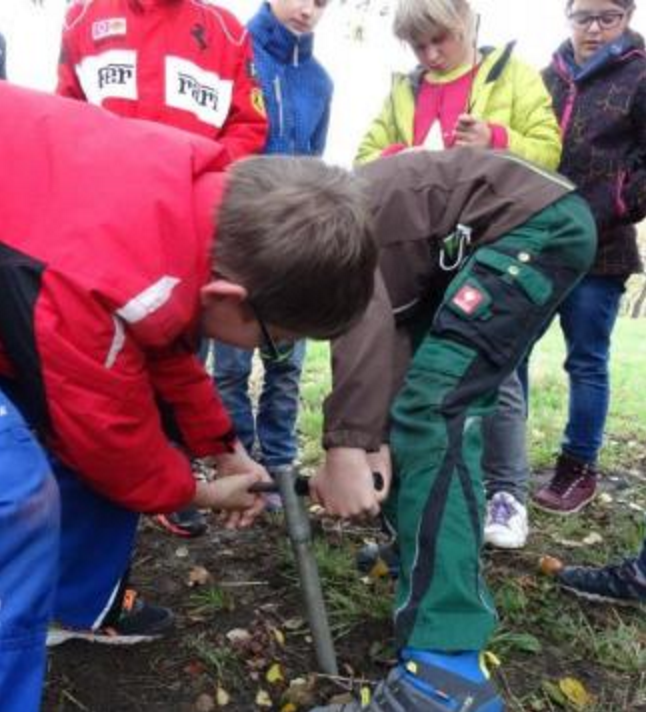 Zwei Kinder  mühen sich nach Kräften, einen Pürckhauer Bohrstock wieder aus dem Boden zu bekommen
