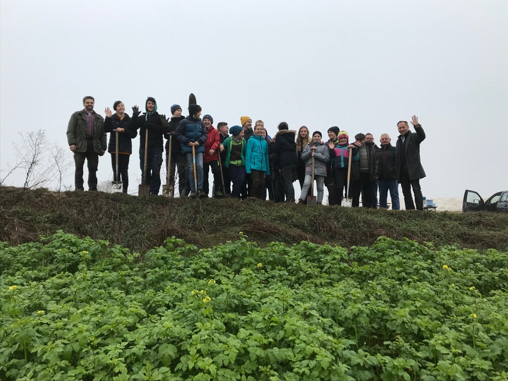 Eine Gruppe Menschen steht auf einem Wegdamm; vom tiefergelegten begrünten Feld im Fordergrund aufgenommen