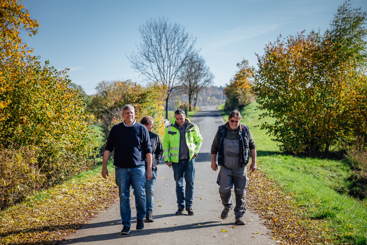 Vier Menschen in Arbeitskleidung (einer in Latzhose, einer mit ALE-Regenjacke) gehen dynamsich einen Feldweg entlang)