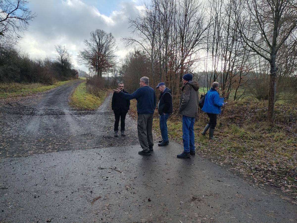 Fünf Männer stehen in der Flur. Im Hintergrund ist eine Weggabelung. An den Rändern der Wege stehen Bäume.