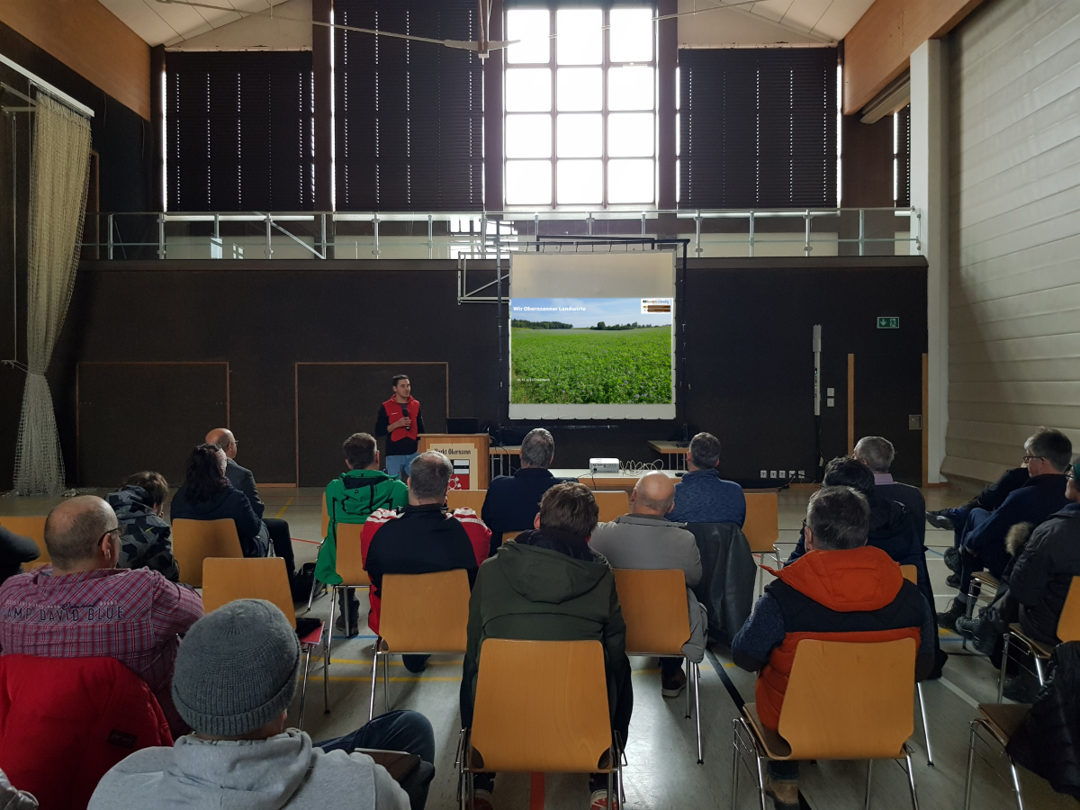 Foto: im Vordergrund sitzen auf Stühlen Menschen, sie schauen auf eine Leinwand im Bildhintergrund, links vor der Leinwand steht ein Landwirt