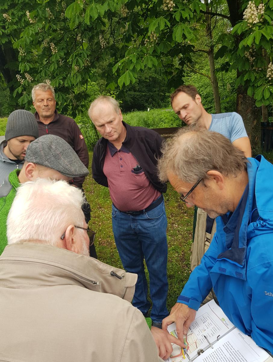 Foto: Eine Gruppe von Menschen beugt sich über einen aufgeschlagenen Ordner mit Blättern mit Text und einer Skizze. Im Hintergrund sind Kastanienblätter.