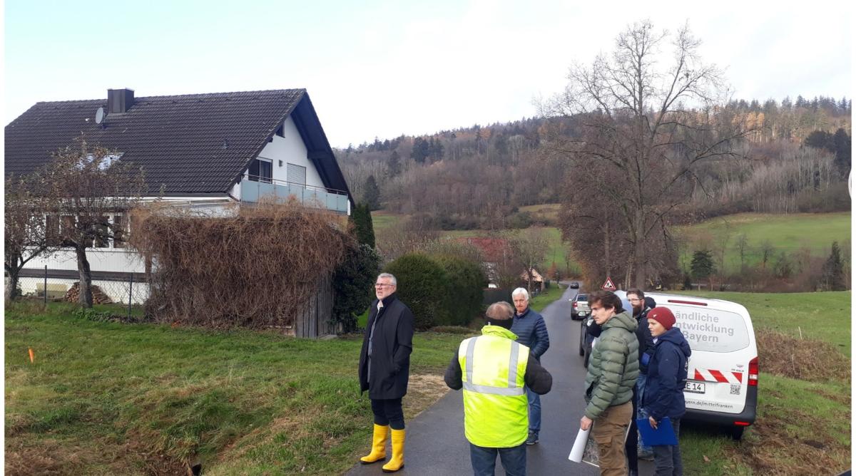 Einige Menschen, z.T. mit Warnweste oder Gummistiefeln, steht vor einem Fahrzeug mit LE-Logo in der Landschaft; am Rand des Bildest ist ein Wohnhaus und eine modellierte Wiesenfläche zu sehen.