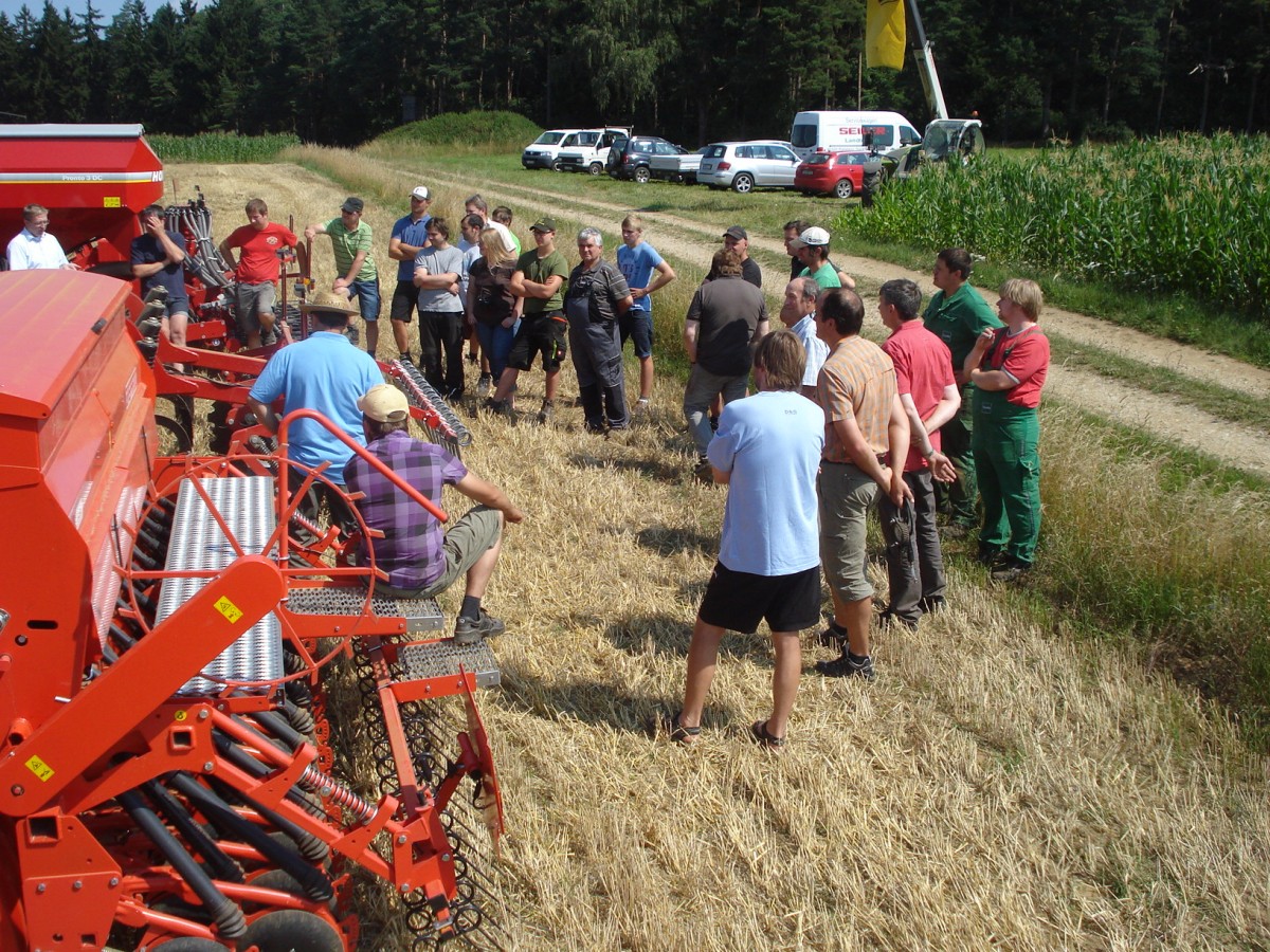 Landwirte lauschen einer Maschinenvorführung auf dem Feld. Manche von ihnen sitzen auf der Drillmaschine im Vordergrund.