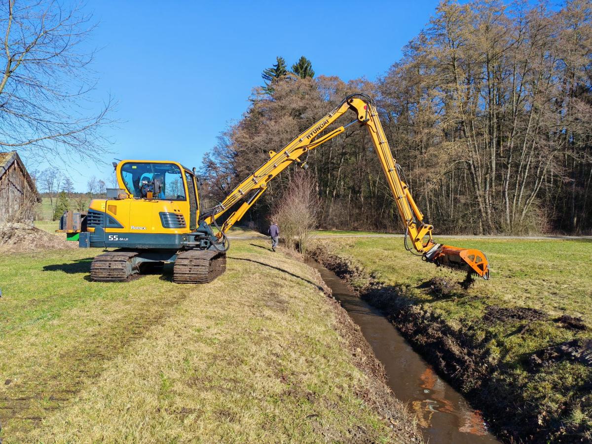 Ein Bagger mit Teleskoparm räumt einen Graben mit einem Mähkorb