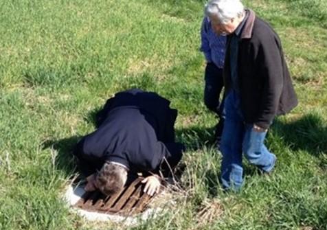 Eine Person kniet vor einem Gulli in der Wiese und schaut hinein