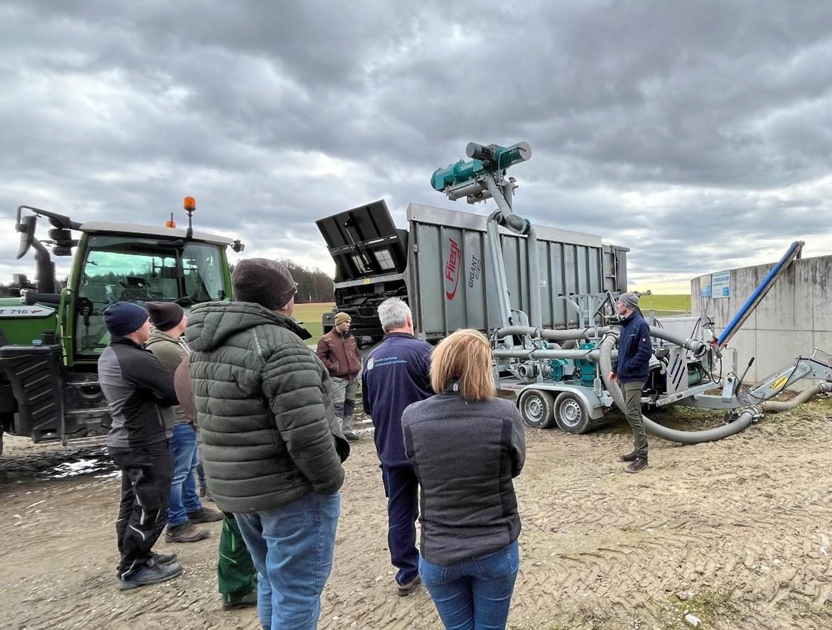 Der Landwirt Florian Huber-Hochradl steht vor einem Traktor mit Autoanhänger, auf dem ein Gülleseparator aufgebaut ist und erläutert die technische Funktionsweise des Separators