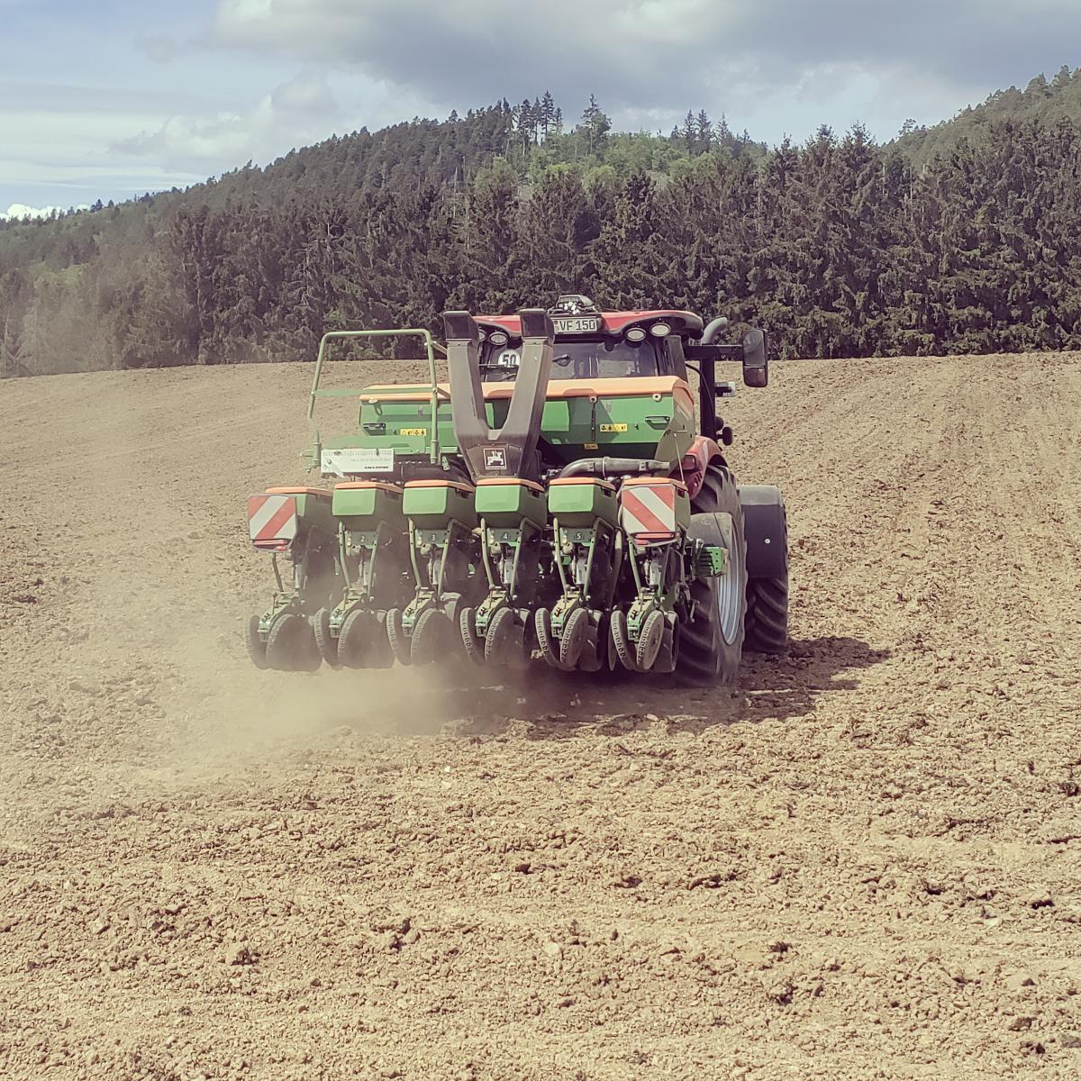 Foto: gegrubbertes Feld mit einem Bulldog und angehängter Sämaschine von hinten, im Hintergrund bewaldete Hügel