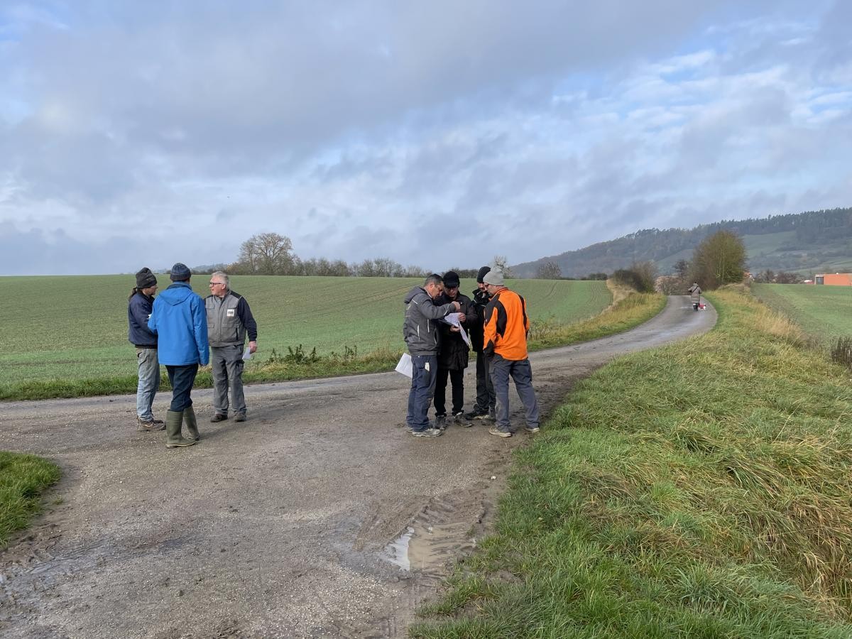 Foto: 2 Gruppen von Männern zu 3 und 4 Personen stehen auf einem Weg, dahinter Acker