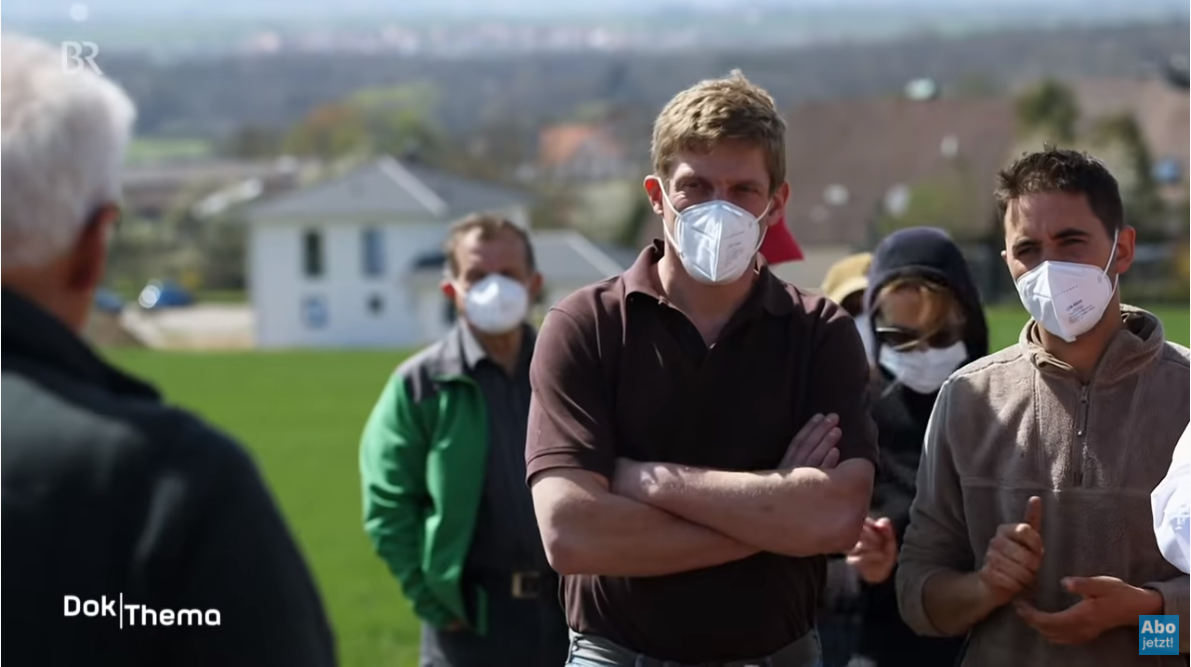 Eine Gruppe Landwirte mit FFP2-Masken blickt entschlossen auf dem boden:ständig-Feldtag in Obernzenn. Am Bildrand sind die Logos des Bayerischen Rundfunk zu sehen, woran man erkennt, dass es sich um einen screenshot aus einer Sendung handelt.