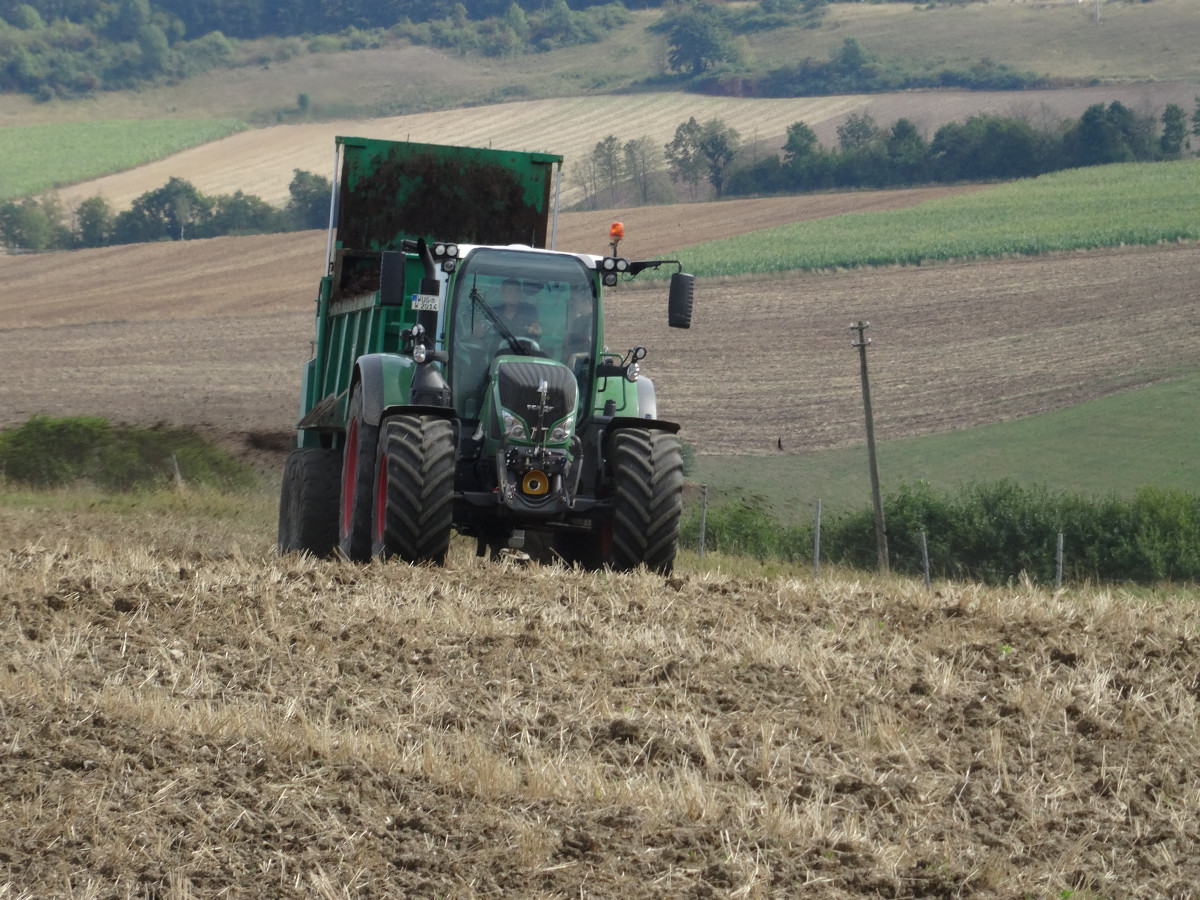 Miststreuer fährt über geneigten, stoppelbearbeiteten Acker in einer hängigen Gegend.