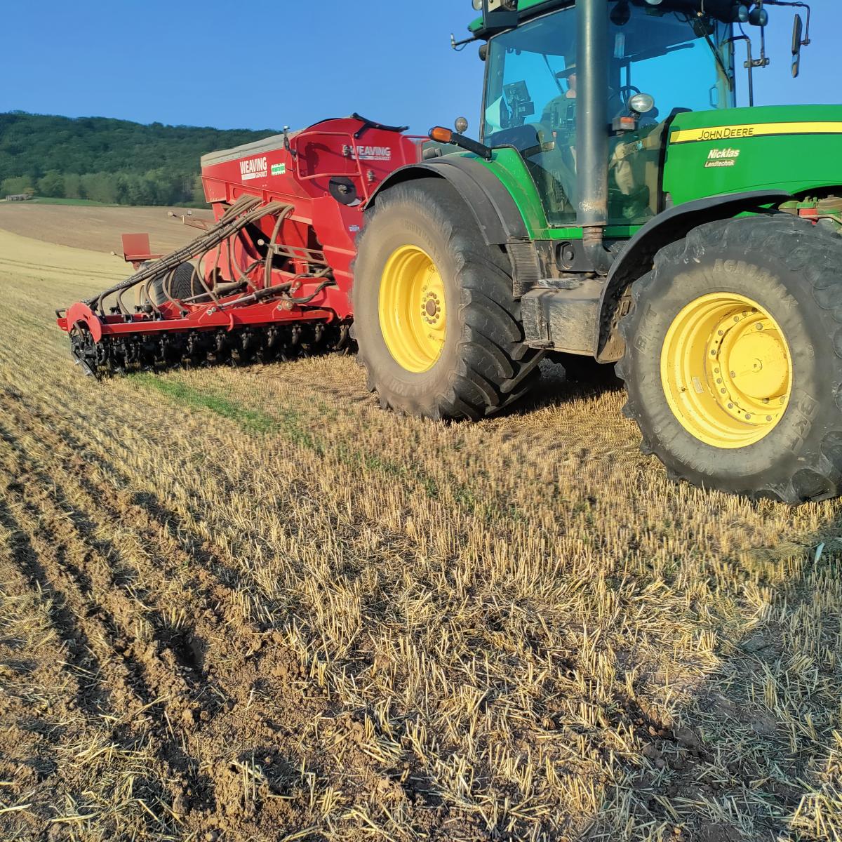 Foto: Stoppelfeld im Vordergrund in Großaufnahme ein grüner Traktor mit Direktsaatmaschine; im Hintergrund ist ein Wald