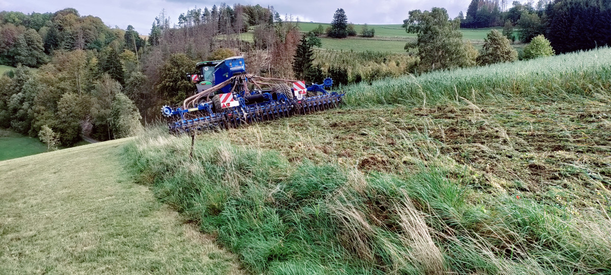 Foto: Mittelgebirgslandschaft, im Vordergrund ein abschüssiges Feld. Ein grüner Traktor zieht eine blaue Direktsaatmaschine vom Betrachter weg. Im Hintergrund Wald und Wiesen.