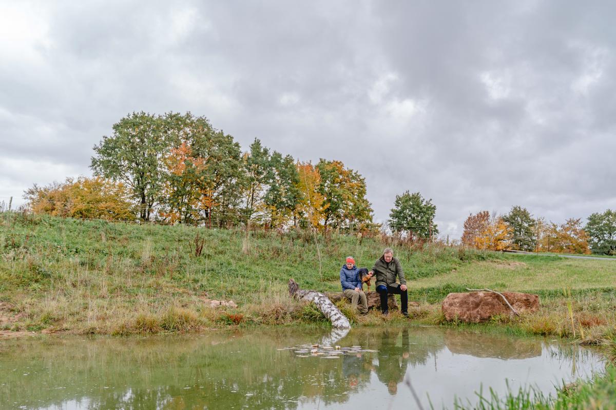 Das Ehepaar Krehan mit Hund sitzt auf einen Stein am Ufer des idyllischen Teichs.