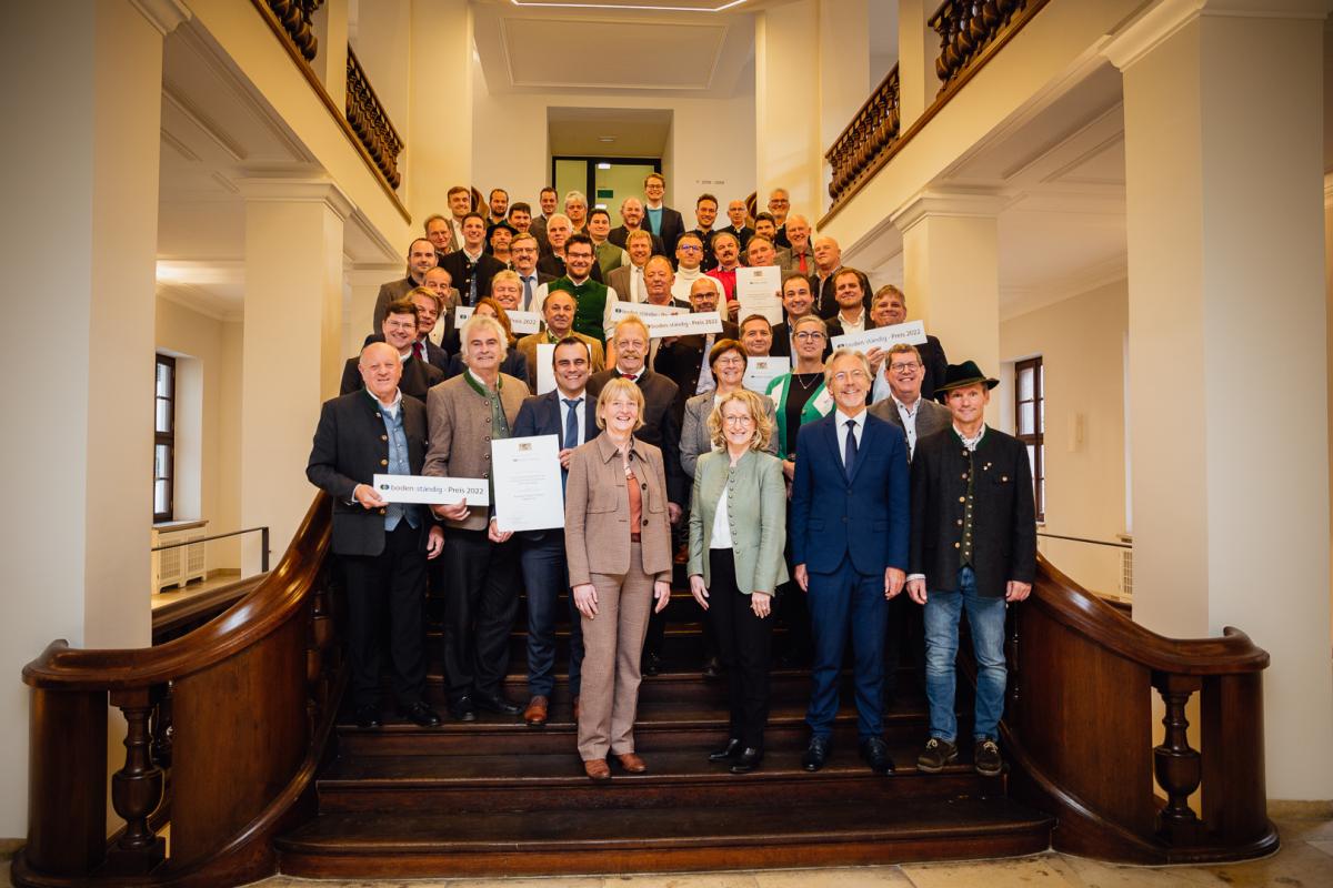 Gruppenbild knapp 50 strahlender Gewinner auf imposanter hölzerner Freitreppe. Manche halten ihre Urkunden resp. die 'boden:ständig-Preis 2022'-Schilder.