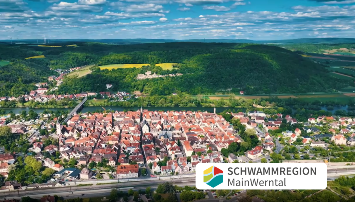 Luftbild mit Siedlung im Vordergrund, dahinter Hügel mit Wald und gelben Feldern, vorne ein Fluss, darüber Wolken. Logo Schwammregion MainWerntal