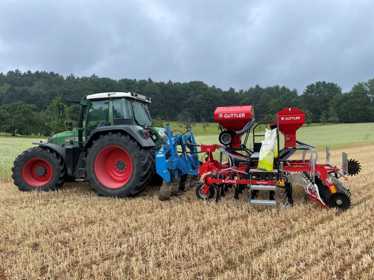 Foto: Auf einem abgeerntetem Feld steht ein Traktor mit einer Direktsaatmaschine, im Hintergrund befinden sich eine Wiese und ein Wald