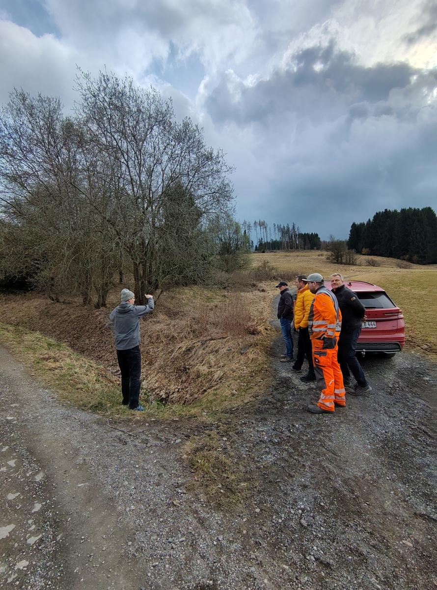 Foto: Ein Mann steht vor einem Graben. Eine Gruppe von 4 Männern schaut in Richtung des Mannes. Im Hintergrund stehen unbelaubte Bäume und ist angefrorenes Günland.