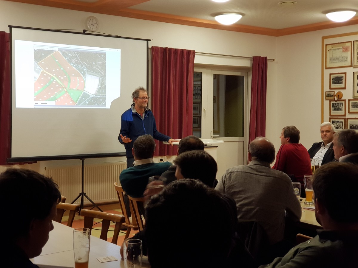 Foto: Raum mit Menschen die an Tischen sitzen, mit dem Rücken zum Betrachter. Im Hintergrund steht ein Mann vor einer Leinwand mit dem Gesicht zum Auditorium