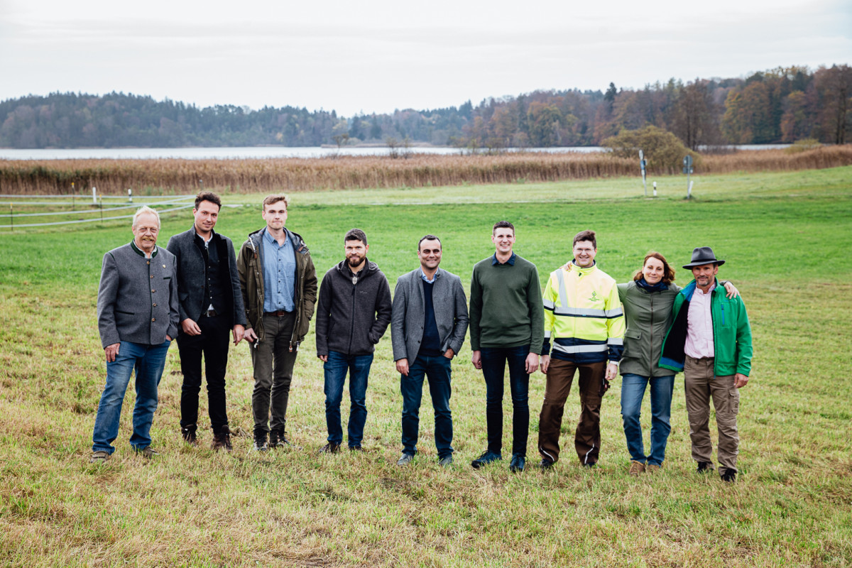 Neun Menschen stehen auf einem Grünlandstreifen - im Hintergrund glänzt hinter einem Schilfgürtel der See.