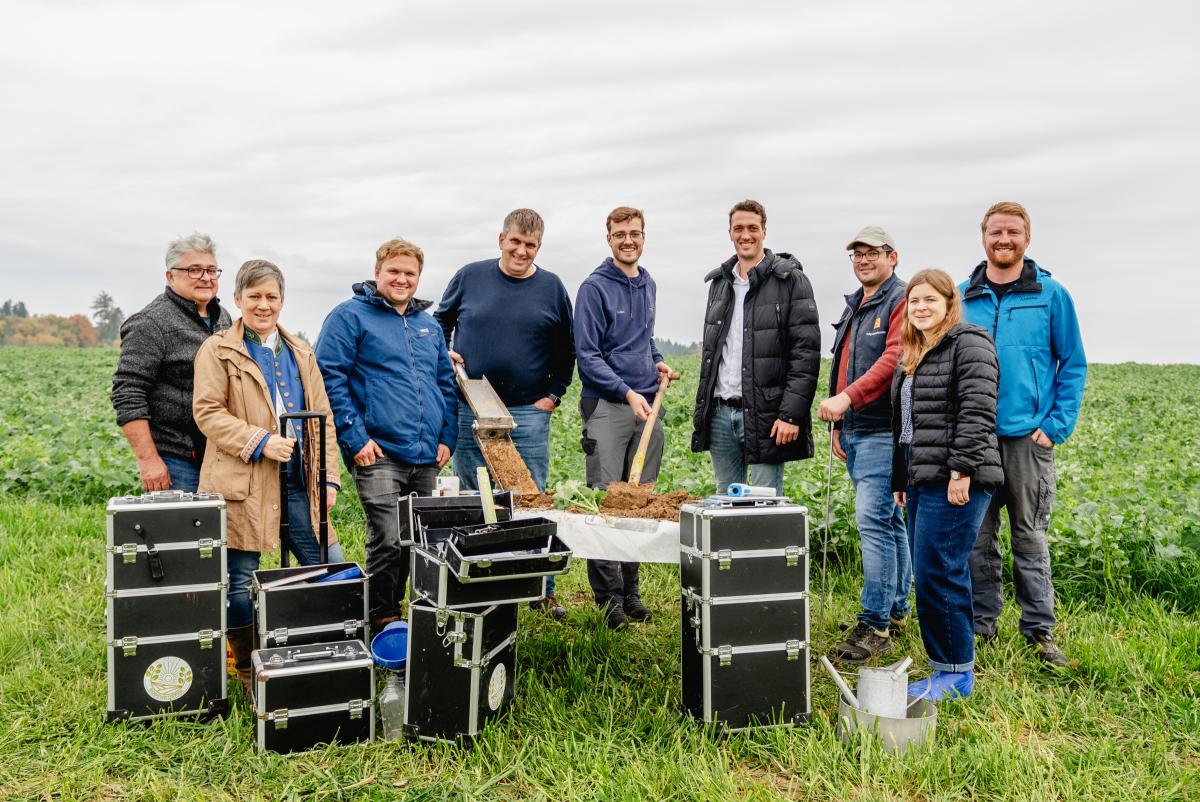 Eine Gruppe Menschen mit großen Werkzeugkoffern und einer Bodenprobe auf einem Tisch am Rande einer Zwischenfruchtfläche