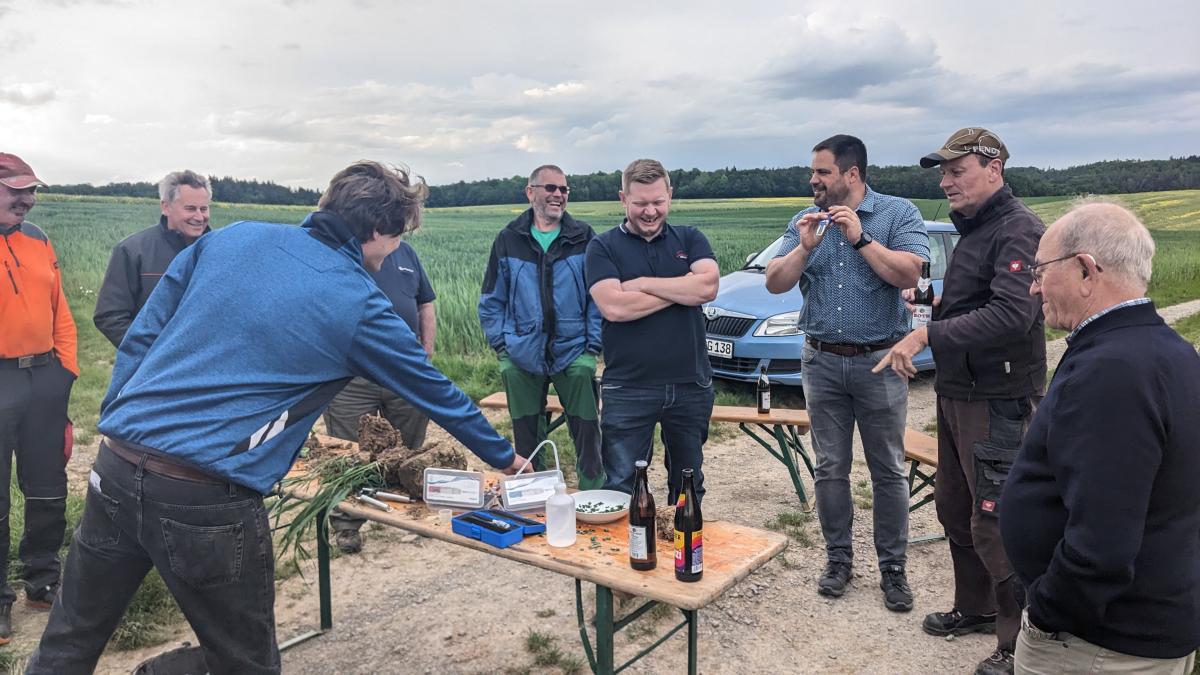 Lachende Landwirte stehen um einen Tisch auf dem Feld, auf dem verschiedene Utensilien zur Bodenuntersuchung aufgebaut sind.