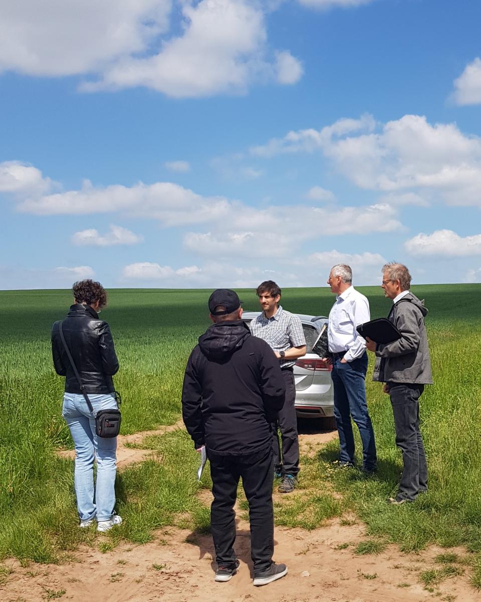 Foto: den Hintergrund bilden Getreidefeld und blauer Himmel mit leichten Wolken, im Vordergrund stehen 5 Personen, dahinter ist das Heck eines Autos