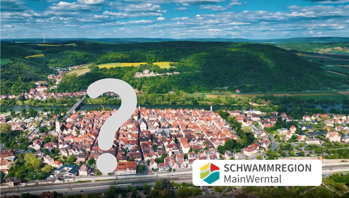 Luftbild mit Siedlung im Vordergrund, dahinter Hügel mit Wald und gelben Feldern, vorne ein Fluss, darüber Wolken. Logo Schwammregion MainWerntal; da wo beim thumpnail die Menschen, ist hier ein Fragezeichen davor.