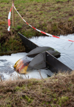 Absperrblech in einem Wiesengraben, in dessen Mitte eine an eine Wippvorrichtung erinnernde Kammer den Durchstrom reguliert. Erkennbar ist der um ca. 15cm höhere Wasserstand stromaufseitig.