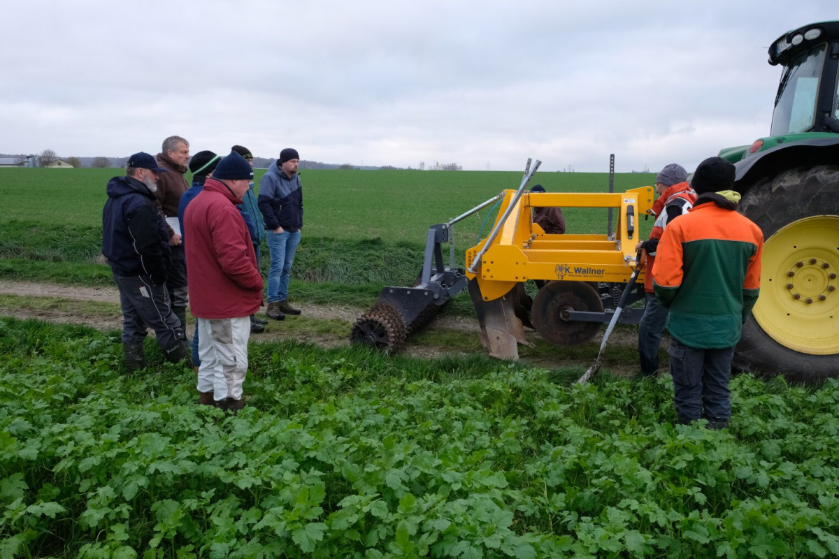 Foto: eine Gruppe von Menschen steht auf einem Feldweg und sieht eine Maschine an die den Boden lockert