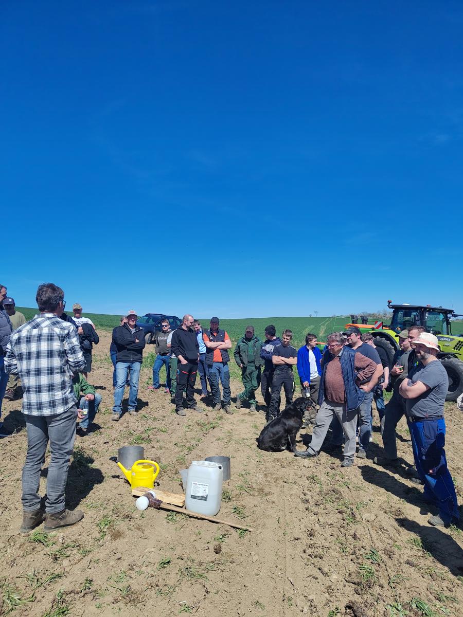 Foto: in der Bildmitte stehen auf einem geackerten Feld um die 20 Personen im Halbkreis um einen Mann im Vordergrund. Vor dem Mann stehen zwei Metallringe mit einem Durchmesser von 40 cm auf dem Feld, im Hintergrund steht ein Bulldog mit einer Sämaschine , dahinter ein grünes Feld und blauer Himmel